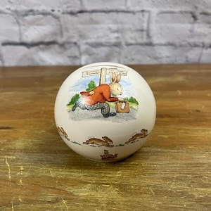 May include: A white ceramic piggy bank shaped like an egg with a Peter Rabbit illustration. The illustration shows Peter Rabbit wearing a red jacket and carrying a suitcase. The signpost reads "Bunny Town" and "To The Station".