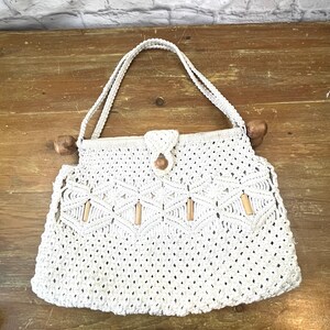 May include: Cream-colored macrame handbag with wooden handles and accents. The bag features a woven design with geometric patterns and a wooden button closure. The handles are made of the same cream-colored material as the bag.