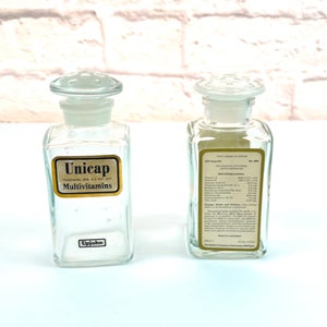 May include: Two vintage glass bottles with gold labels. The bottle on the left says "Unicap Multivitamins" and "Upjohn". The bottle on the right has a list of vitamins and minerals, and says "The Upjohn Company, Kalamazoo, Michigan".