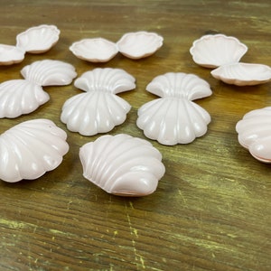 May include: A collection of light pink seashell-shaped decorative items. The shells vary in size and design, with some appearing open and others closed. The items are arranged on a wooden surface, creating a beach or ocean-themed aesthetic.