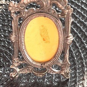 May include: An antique silver-toned picture frame with an oval aperture. The frame features decorative floral and scrollwork details. The interior of the frame has a yellow-orange background. The frame is set against a dark, textured surface.