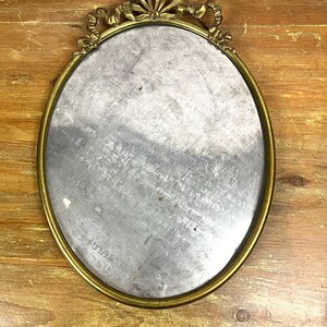 May include: An oval-shaped brass mirror with a decorative, ornate frame featuring a bow design. The mirror has a silver-toned reflective surface.