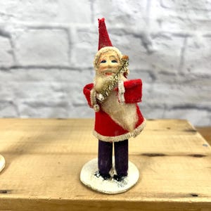 May include: A vintage cotton-stuffed Santa Claus figurine with a red and white suit, a white beard, and a red hat. He is holding a gold and white tinsel stick in his right hand. The figurine is standing on a white base.