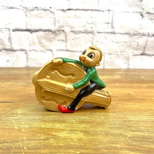 Op de afbeelding: Een keramiek beeldje van een elf gekleed in groen die een groot, bruin muziekinstrument bespeelt. De elf heeft een grillige uitdrukking en houdt het instrument vast met beide handen.