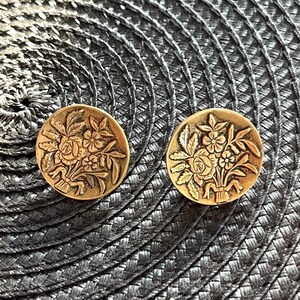 May include: Two antique gold-colored round buttons. Each button features a detailed floral design with roses, leaves, and other flowers. The buttons are set against a textured, dark gray background.