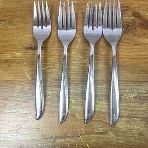 Puede incluir: Cuatro tenedores de acero inoxidable con un diseño de estrella en el mango. Los tenedores están dispuestos verticalmente sobre una superficie de madera, mostrando sus púas y mangos delgados. Los tenedores miden aproximadamente 20 cm de largo.