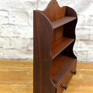 May include: A small, dark brown wooden shelf with two shelves and a drawer. The shelf has a decorative top with a curved design and two small wooden knobs on the drawer. The wood has a rich, warm tone.
