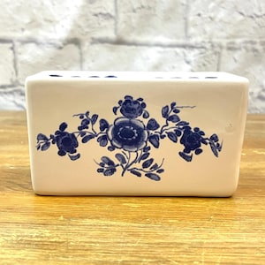 May include: A rectangular, off-white ceramic toothbrush holder with a blue floral design. The holder has multiple holes on top for toothbrushes and features a symmetrical floral pattern in the center. The background is a wooden surface and a white brick wall.