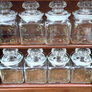 May include: A wooden spice rack with eight clear glass jars. Each jar has a clear lid and is decorated with a green leaf pattern. The jars are arranged in two rows of four.