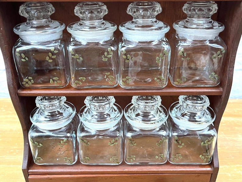 May include: A wooden spice rack with eight clear glass jars. Each jar has a clear lid and is decorated with a green leaf pattern. The jars are arranged in two rows of four.