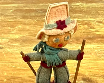 Puede incluir: Una muñeca de fieltro con un sombrero rosa con una estrella roja, una bufanda azul y pantalones grises. La muñeca sostiene dos palos de madera y lleva botas de fieltro rojas.