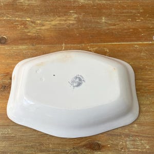 May include: A white, rectangular ceramic serving dish with rounded edges. The dish has a slightly raised rim and a faded, grey emblem in the centre. The dish is set against a wooden surface.