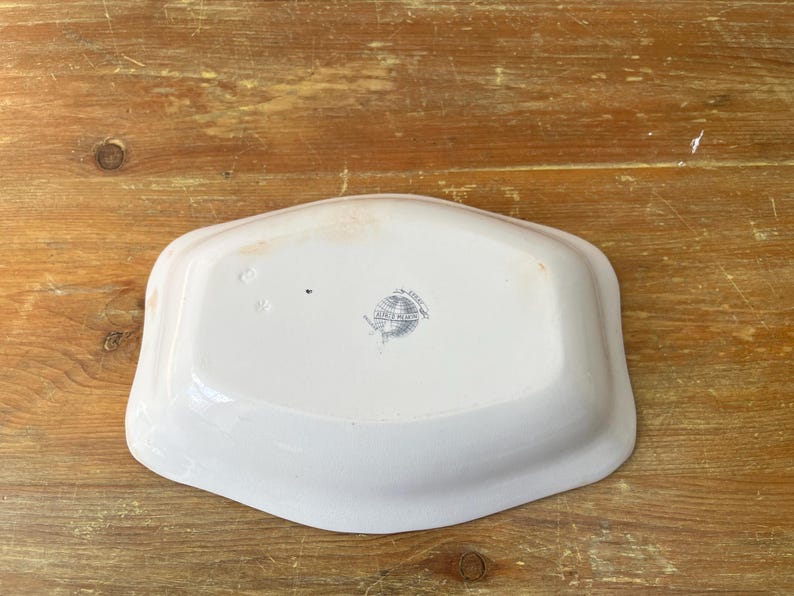 May include: A white, rectangular ceramic serving dish with rounded edges. The dish has a slightly raised rim and a faded, grey emblem in the centre. The dish is set against a wooden surface.
