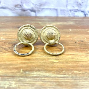 May include: Two antique-style cabinet pulls with a distressed, aged appearance. The pulls are circular with a decorative design and a ring handle. The color is a muted gold, and they are set against a wooden surface.