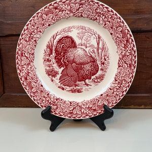 Puede incluir: Un plato de cena de cerámica blanca con un diseño rojo y blanco de un pavo en un entorno de bosque. El plato tiene un borde decorativo de hojas y vides rojas.