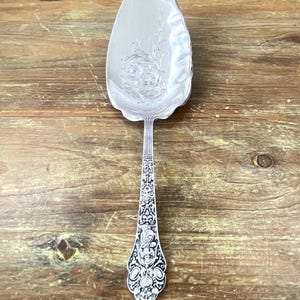 May include: A silver pie server with an ornate handle and a wide, leaf-shaped blade. The blade features an embossed floral design. The handle is intricately detailed with scrolling patterns and decorative elements. The server rests on a wooden surface.