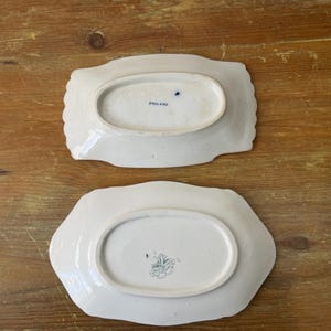 May include: Two white ceramic serving dishes with scalloped edges. The dishes have an oval centre and are stamped with text and a design on the base. One dish is stamped "ENGLAND".