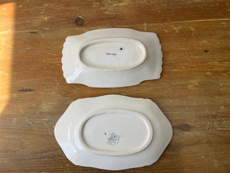May include: Two white ceramic serving dishes with scalloped edges. The dishes have an oval centre and are stamped with text and a design on the base. One dish is stamped "ENGLAND".