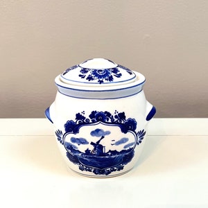 Może przedstawiać: Biały ceramiczny dzbanek z niebiesko-białym wzorem Delft. Dzbanek ma pokrywkę i przedstawia scenę z wiatrakiem z niebieskimi i białymi kwiatowymi akcentami.