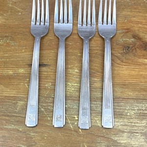 May include: Four silver forks with tines and ridged handles. Each fork has a decorative floral design at the base of the handle. The forks are arranged on a wooden surface.
