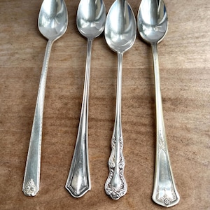 Könnte beinhalten: Vier silberne Löffel mit unterschiedlichen Griffmustern werden auf einer braunen Holzoberfläche präsentiert. Die Löffel haben verschiedene Griffstile, darunter verzierte Blumenmuster und einfache, geometrische Formen. Die Löffel sind glänzend.