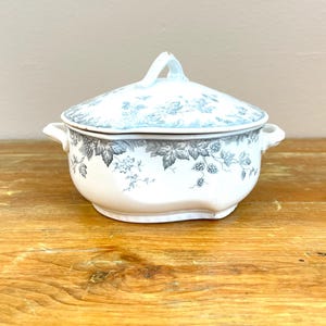 May include: A white ceramic tureen with a lid, featuring a gray botanical pattern of leaves and hop-like clusters. The vessel has two side handles and a central handle on the lid, with a scalloped rim and a rounded, slightly oval base shape.