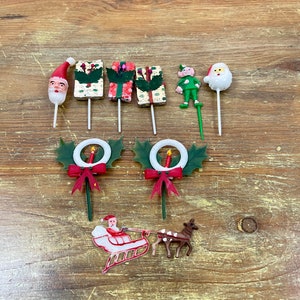 May include: A collection of vintage Christmas decorations on wooden surface. Includes Santa Claus, an elf, gift boxes, wreaths, and a Santa sleigh with a reindeer. The decorations are in various colors, including red, green, and white.