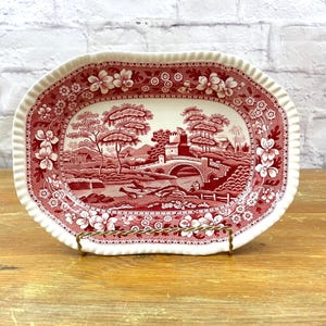 May include: An oval ceramic serving dish featuring a deep red transferware pattern on a cream background. The central scene depicts a stone bridge, trees, and buildings, while the scalloped rim is decorated with a detailed floral border.