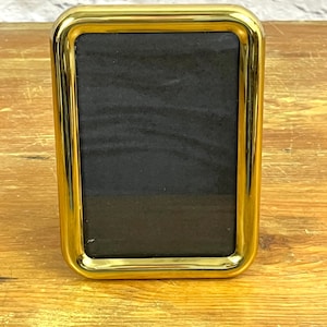 May include: A rectangular gold-colored picture frame with a black interior. The frame has rounded corners and a glossy finish. The frame is sitting on a wooden surface.