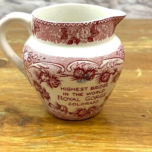 Puede incluir: Una pequeña jarra de cerámica blanca con asa y boquilla. La jarra está decorada con un patrón floral y paisajístico rojo. El texto en la jarra dice "HIGHEST BRIDGE IN THE WORLD ROYAL GORGE COLORADO."
