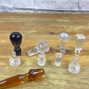 May include: A collection of eight glass bottle stoppers, including a black glass stopper, a clear glass stopper with a textured surface, a clear glass stopper with a round top, a clear glass stopper with a square top, and a clear glass stopper with a round top and a textured surface. The stoppers are arranged on a wooden surface.