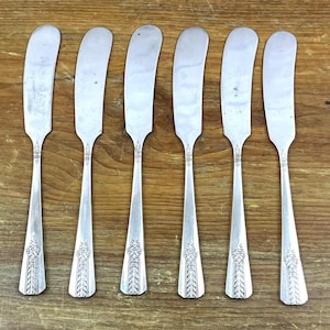 May include: Six silver butter knives with curved blades and ornate floral designs on the handles are arranged on a wooden surface. The knives have a vintage aesthetic, suggesting they are antique or vintage.