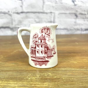May include: A small white creamer with a red and white design of a building with trees and a boat. The creamer has a handle and a spout.