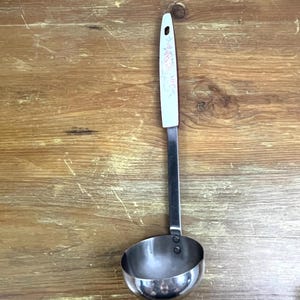 May include: A silver ladle with a white handle featuring a floral design. The handle is attached to the ladle with a silver metal band.