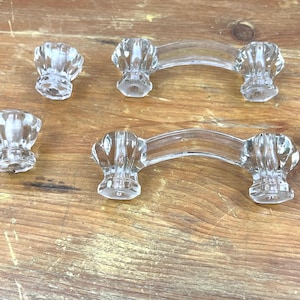 Könnte beinhalten: Schrankknöpfe und -griffe aus klarem Glas. Die Knöpfe haben ein abgerundetes, geriffeltes Design. Zwei Griffe haben ein gebogenes Design mit den gleichen geriffelten Enden. Die Hardware wird auf einer Holzoberfläche ausgestellt.