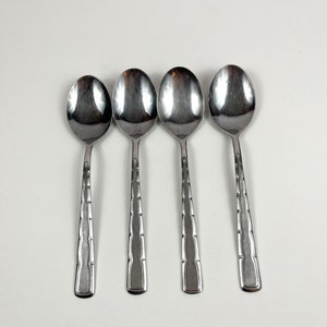 May include: Four silver spoons with a decorative pattern on the handle. The spoons are arranged in a row on a white surface.