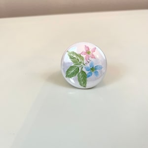 May include: White ceramic knob with a painted design of pink and blue flowers with green leaves. The knob has the text "N.B.S." written on it.