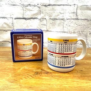 May include: A white ceramic 1994 calendar mug with a handle, displayed next to its original box. The mug features a calendar design with yellow, red, and blue stripes. The box is dark blue with the text "1994 Calendar Mug".