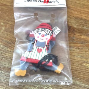 May include: A wooden ornament in a clear plastic bag labeled Larsen Danmark A/S. The ornament depicts a figure wearing a red hat, blue clothing, and a white apron with green stripes and a red heart. The figure holds a sign reading GOD JUL.