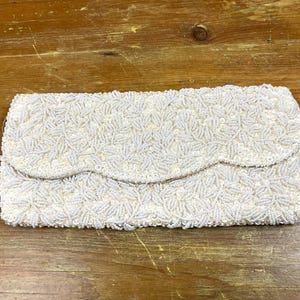 May include: A vintage white beaded clutch with a scalloped flap closure. The rectangular bag is covered in small, white beads, creating a textured surface. The clutch is set against a wooden background.