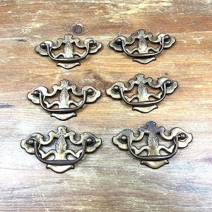 May include: Six antique brass drawer pulls with ornate designs. Each pull features a curved handle and decorative backplate. The pulls are arranged on a wooden surface, showcasing their aged patina and intricate details. Ideal for vintage furniture restoration.