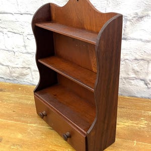 May include: A small, dark brown wooden shelf with two shelves and a drawer. The shelf has a decorative, scalloped top and two small wooden knobs on the drawer. The wood grain is visible, and the shelf appears to be vintage.