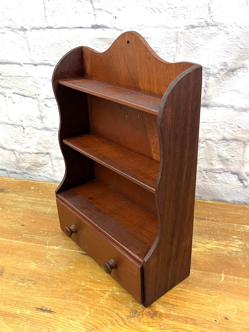 May include: A small, dark brown wooden shelf with two shelves and a drawer. The shelf has a decorative, scalloped top and two small wooden knobs on the drawer. The wood grain is visible, and the shelf appears to be vintage.