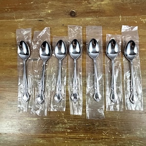 May include: Seven silver teaspoons in individual plastic packaging. The spoons have ornate handles and are arranged in a row.