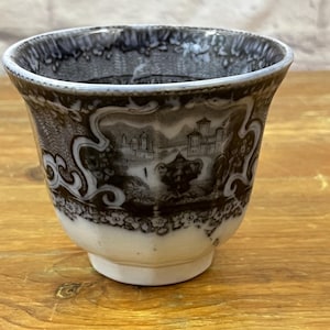 May include: A vintage teacup with a black and white landscape design. The cup has a flared shape with a white base and a detailed scene of a castle and trees. The rim has a decorative pattern.