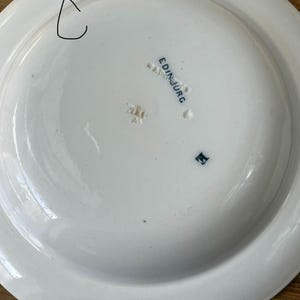 May include: A white ceramic plate with a wide rim. The underside features the word "EDINBURG" in blue lettering, along with other markings. The plate has a small chip on the rim.