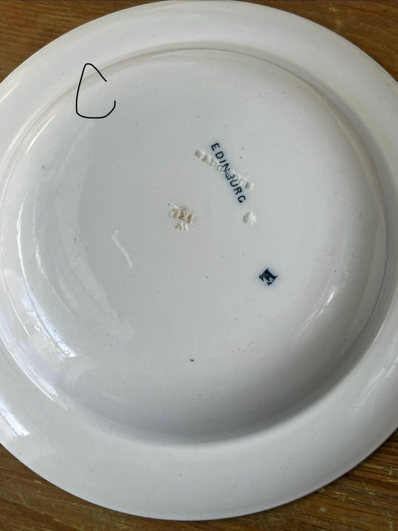 May include: A white ceramic plate with a wide rim. The underside features the word "EDINBURG" in blue lettering, along with other markings. The plate has a small chip on the rim.