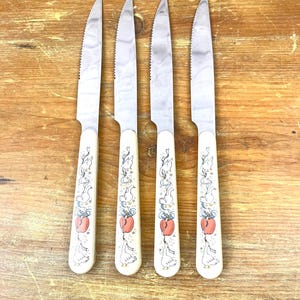 May include: A set of four stainless steel serrated knives with off-white handles. Each handle features a decorative illustration of white geese, blue ribbons, and a red apple. The knives are arranged vertically in a parallel row on a wooden surface.