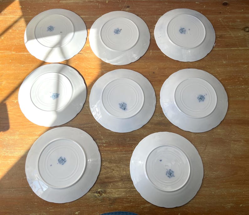 May include: Eight white ceramic plates with scalloped edges are arranged on a wooden surface. Each plate has a blue floral design in the centre. The plates are viewed from the underside.