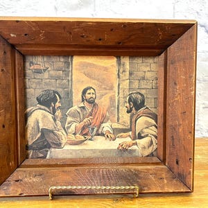 Op de afbeelding: Een ingelijste prent die een religieuze scène toont met figuren rond een tafel. De prent staat in een rustieke houten lijst met een messing ezelrug. Het kunstwerk gebruikt gedempte tonen en heeft een historisch thema.